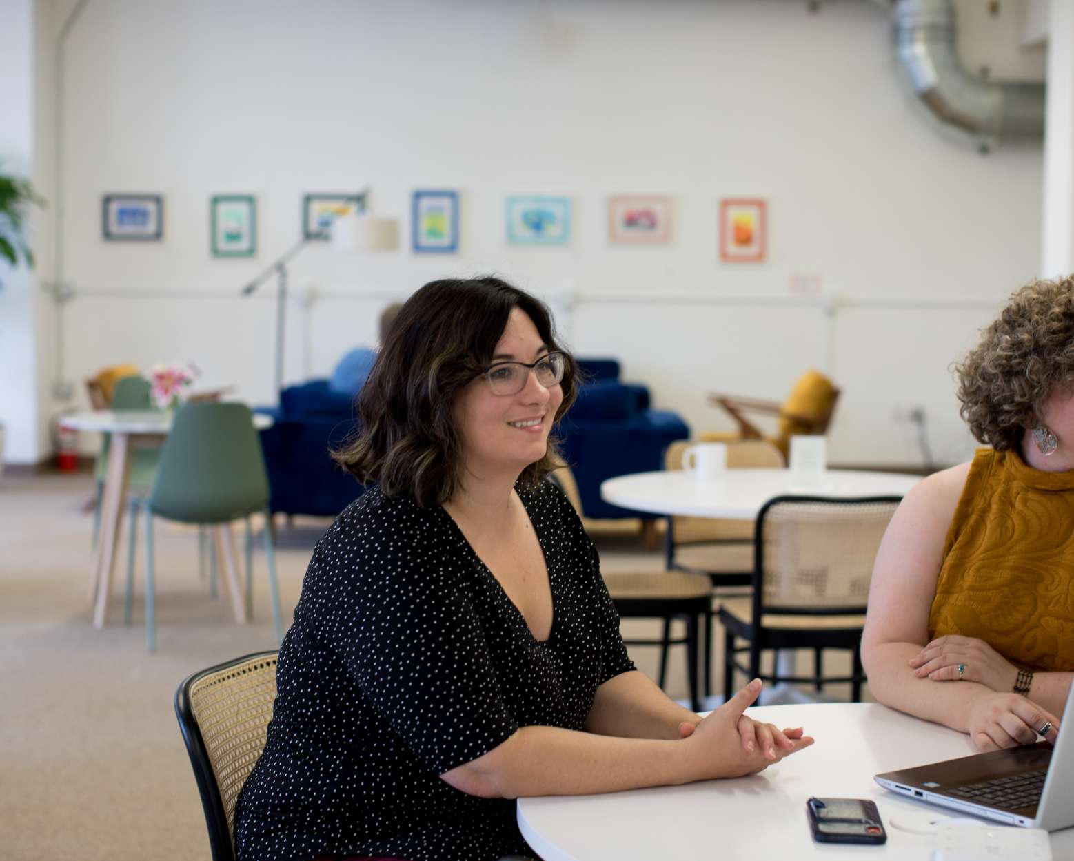 Two feminine people sit next to each other at a table with a laptop, smiling.