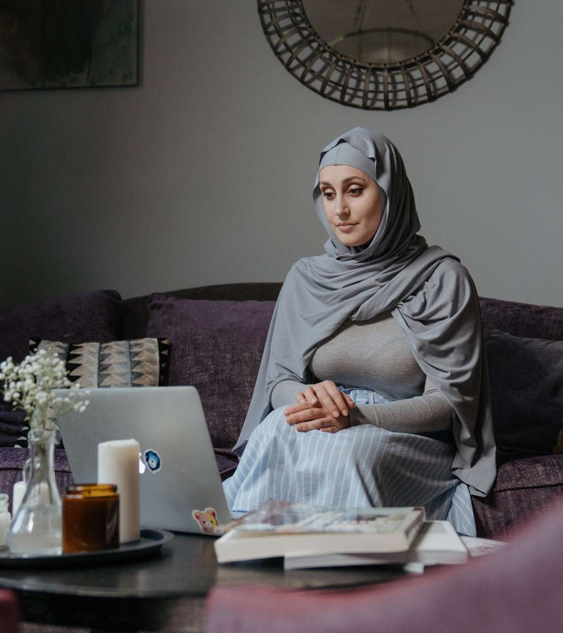 Woman with fair skin wearing a hijab sits on a couch and looks at an open laptop.