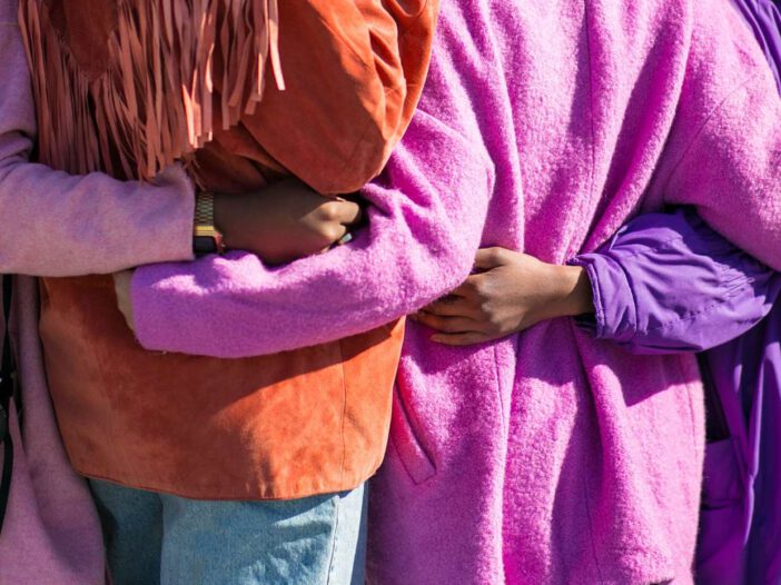 The backs of four people with brightly colored coats and blue jeans with their arms wrapped around each other's waists.