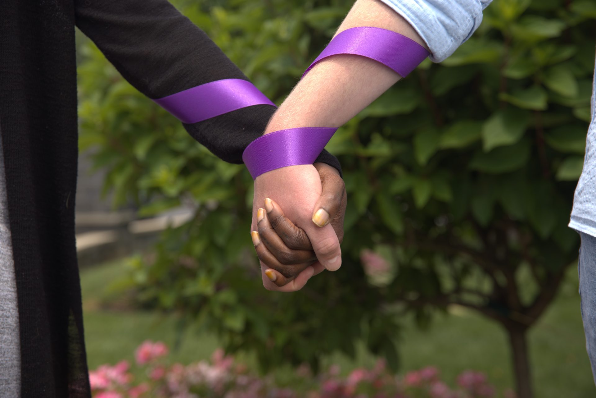 Two people with different skin tones hold hands outside, their joined hands wrapped in purple ribbons.